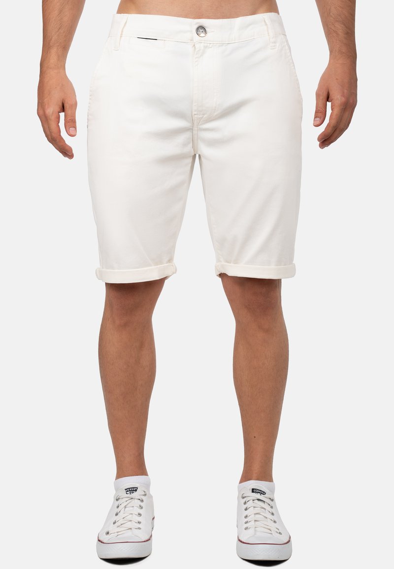 Witte katoenen shorts met opgerolde uiteinden, voorzien van een knoopsluiting en zijzakken. Draagt witte sneakers op een neutrale achtergrond.