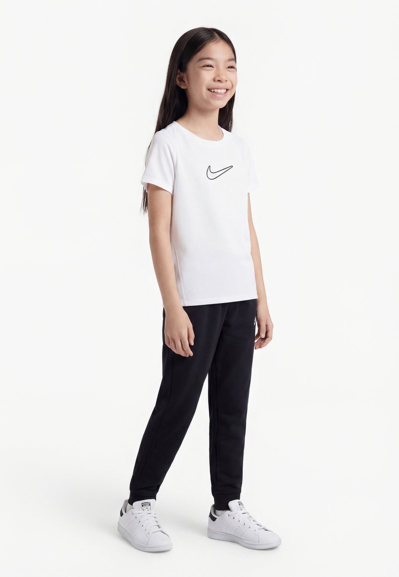 Nasmejana deklica stoji oblečena v belo majico Nike, črne hlače in bele superge na enobarvnem belem ozadju.