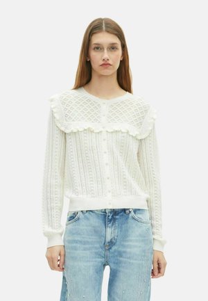 Jonge vrouw in een witte textuurgebreide cardigan met ruches en blauwe jeans, staand tegen een effen witte achtergrond.