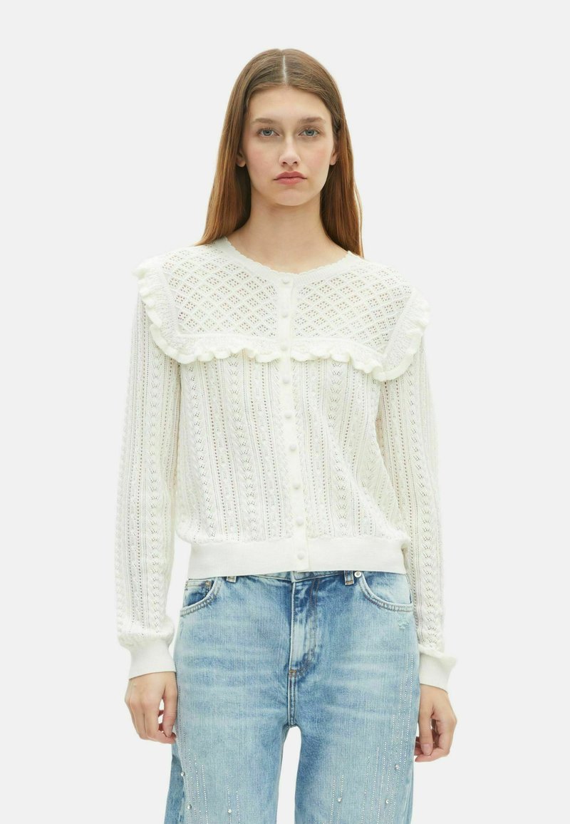 Giovane donna che indossa un cardigan in maglia bianca con dettagli arricciati e jeans blu, in piedi su uno sfondo bianco semplice.