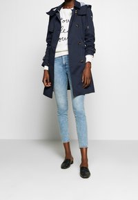 Osoba w granatowym płaszczu, białym swetrze z czarnym napisem, jasnoniebieskich obcisłych dżinsach i czarnych butach slip-on na tle białej ściany.