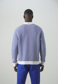 Sininen ja valkoinen raidallinen neule, jossa on valkoinen kaulus, taitettavat hihansuut ja helma, valmistettu pehmeästä materiaalista. Päällä siniset housut.