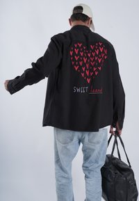 Črna majica z rdečim vzorcem srčkov na hrbtni strani, z besedilom "SWEET heart." Oblečena preko svetlo modrih džinsov, z črno torbico v roki.