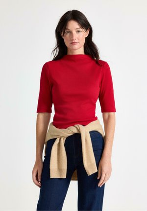 Donna con capelli scuri che indossa una maglietta rossa aderente e jeans scuri con un maglione beige legato intorno alla vita su uno sfondo semplice.