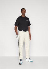 Μαύρο βαμβακερό t-shirt, κρεμ παντελόνι, λευκά γκολφ παπούτσια με μπλε λεπτομέρειες. Κρατώντας ένα γκολφ μπαστούνι, φοράει λευκό γάντι, στέκεται μπροστά σε γκρι φόντο.