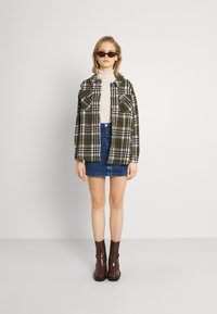 Veste à carreaux verte et blanche avec deux poches poitrine, col roulé beige, mini-jupe en denim bleu et bottines marron.