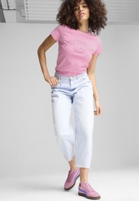 Růžové tričko s krátkým rukávem a nápisem "SPORTSWEAR", světle modré džíny s vysokým pasem a růžové tenisky s fialovými akcenty a hnědými podrážkami.
