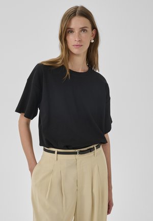 Vrouw met lichtbruin haar, zilveren oorbellen en een losse zwarte korte mouw blouse, gestopt in beige geplooide broek met een zwarte riem.