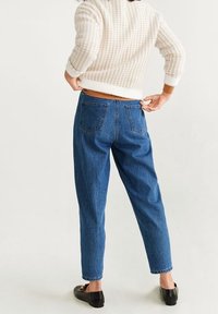 Blaue hoch geschnittene Jeans mit lockerer Passform, ausgestattet mit zwei Gesäßtaschen und einem hellen Waschlook. Kombiniert mit einem cremefarbenen strukturierten Pullover.