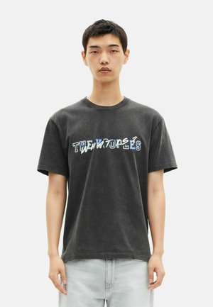 Zwarte katoenen t-shirt met de tekst "DE NEOTROPIE" in meerkleurige vervormde letters, met een relaxte pasvorm en korte mouwen.