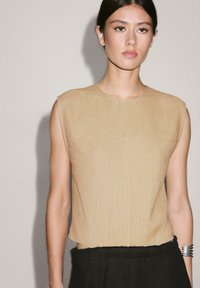 Beige ärmelloses Oberteil aus strukturiertem Stoff, Rundhals und leicht verkürztem Schnitt, kombiniert mit einem dunklen schwarzen Outfit.