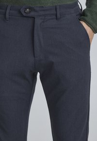 Marinblå byxor i texturerad tyg med knappstängning, fickor med sprund och skräddarsydd passform. Detalj på midjebandet synlig.