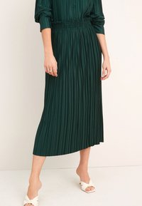 Jupe midi plissée verte avec une taille froncée. Tissu lisse, design fluide et une fente sur le côté. Portée avec des talons ouverts crème.