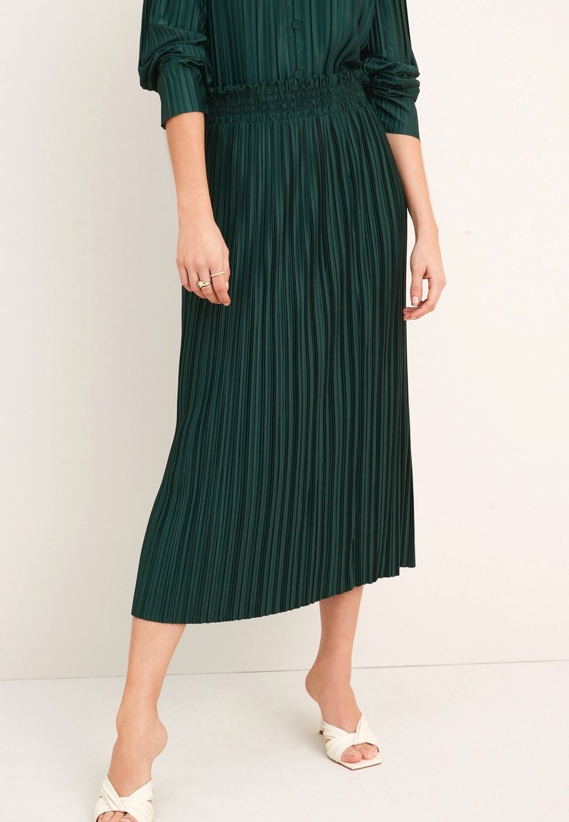 Jupe midi plissée verte avec une taille froncée. Tissu lisse, design fluide et une fente sur le côté. Portée avec des talons ouverts crème.
