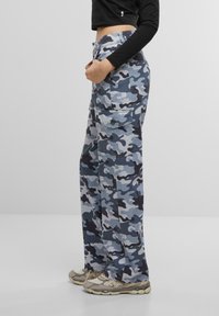 Pantalones cargo en un patrón de camuflaje azul y gris con bolsillos laterales, un corte holgado y cierre de botón en la cintura. El material parece resistente.