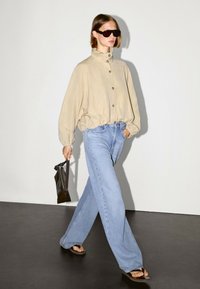 Beżowa, oversizeowa koszula na guziki z wysokim kołnierzem, jasnoniebieskie jeansy o szerokich nogawkach i czarne okulary przeciwsłoneczne, trzymająca czarną kopertówkę w jednej ręce.