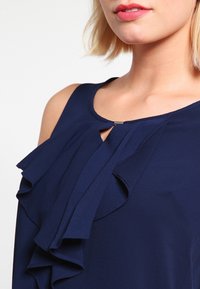 Granatowa bluzka z falbankami na ramionach, dekoltem w kształcie łezki oraz gładką, lekką tkaniną.