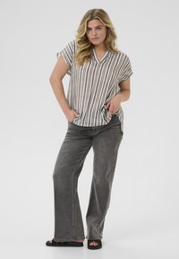 Haut à manches courtes rayé en beige et gris, décolleté en V, assorti à un jeané gris évasé et des sandales noires, debout avec les mains dans les poches.