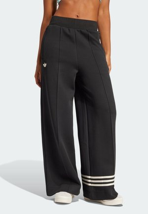 Vrouw draagt zwarte wijde broek met zijzakken, Adidas-logo op de heup, drie witte strepen bij de zoom en witte sneakers.