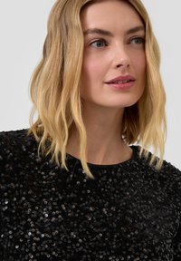 Zwart pailletten top met een ronde hals, glinsterende textuur en korte mouwen, gepresenteerd tegen een neutrale achtergrond.