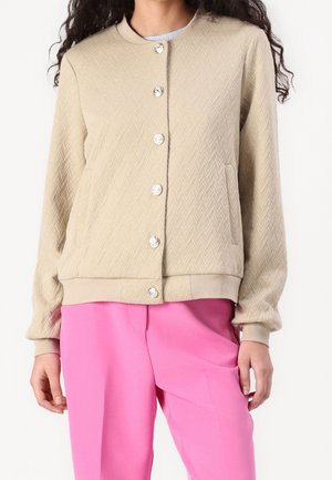 Beige geweven blousejack met geribbelde manchetten en zoom, gecombineerd met felroze broek, getoond op een persoon van nek tot heupen.