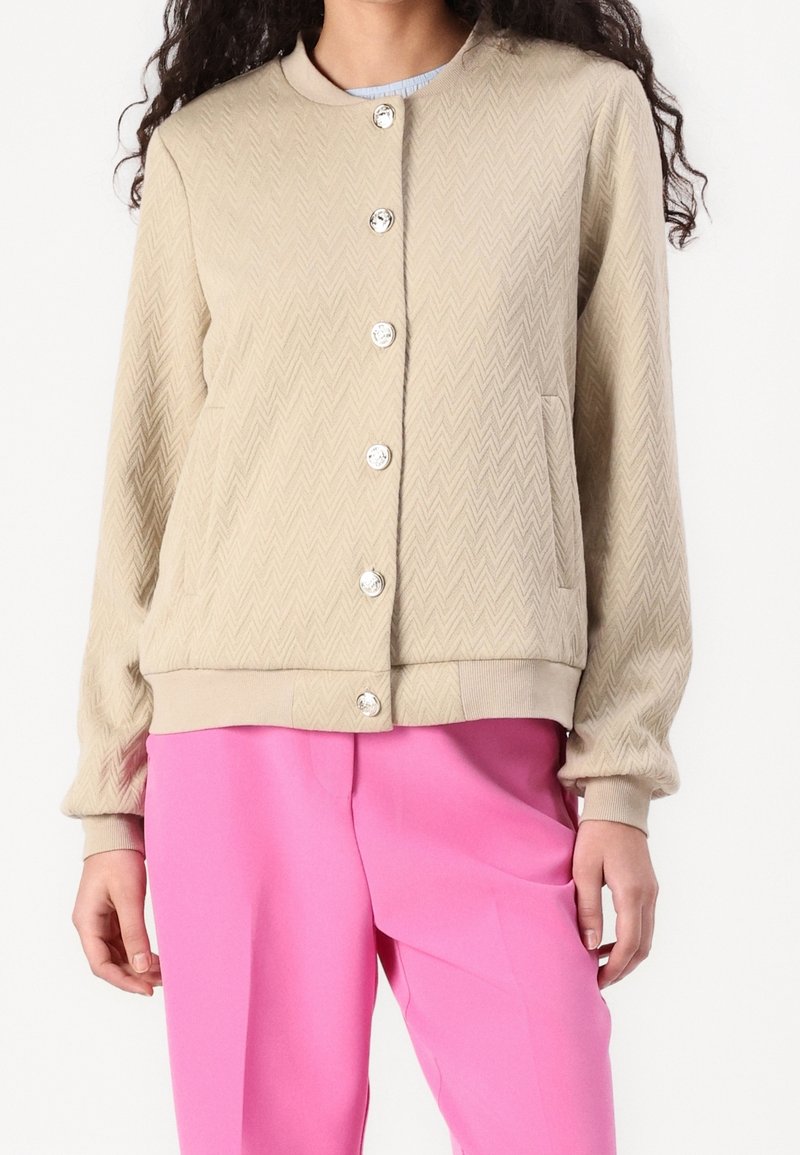 Veste boutonnée beige texturée avec poignets et ourlet côtelés, associée à un pantalon rose vif, portée par une personne visible du cou aux hanches.