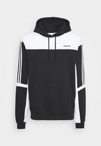 Svart och vit hoodie med en framsida ficka, vita ärmar och svarta tre-stripes accenter. Materialet verkar mjukt och texturerat.