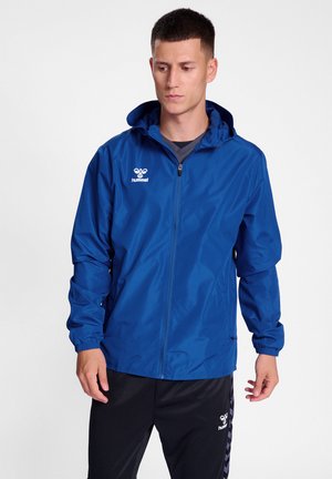 Blauwe lichte jas met een rits aan de voorkant, verstelbare capuchon, elastische manchetten en zijzakken. Heeft een klein wit logo op de borst.