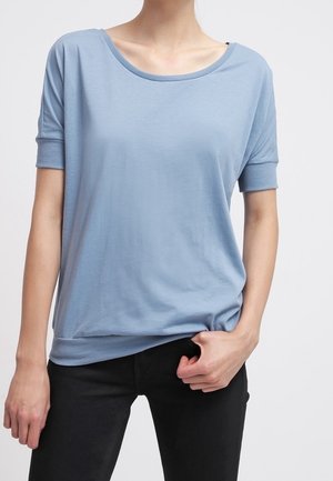 Woman wearing a light blue short-sleeve top with ribbed cuffs, paired with black pants, posing with one hand holding the hem.