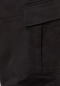 Gros plan sur un tissu noir avec une grande poche rectangulaire et un détail de rabat cousu en haut à droite.
