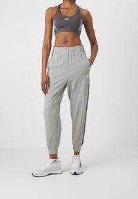 Athletic person wearing gray Adidas sports bra, light gray Adidas joggers with stripes, and white Adidas running shoes standing with hands in pockets.