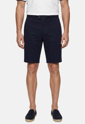 Mann trägt dunkelblaue knielange Shorts, weißes Kurzarmhemd und dunkle Loafers, steht mit entspannten Händen an den Seiten.