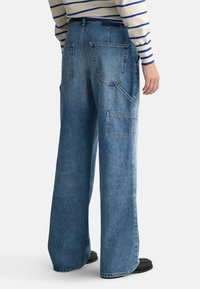 Breda jeans i blå denim med stora cargofickor, en avslappnad passform och en urblekt yta. Synliga sömdetaljer på fickorna.