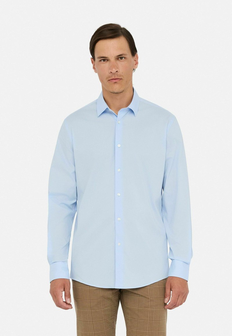 Camisa de manga larga azul claro con botones en el frente, cuello clásico y textura suave, combinada con pantalones de cuadros marrones.