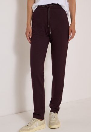 Personne portant un pantalon droit violet foncé avec une taille à cordon et des baskets beiges, debout devant un fond clair uni.