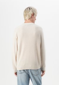 Beige knitted sweater with a textured pattern, long sleeves, ribbed cuffs and hem, worn over light blue jeans. Back view visible.
