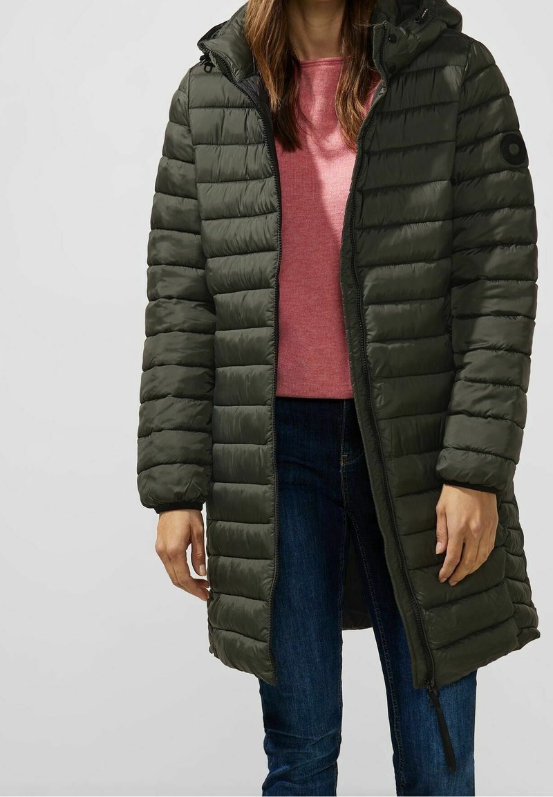 Olivegrön puffjacka med horisontell quiltning, framdragkedja och huva. Bärs över en rosa topp med blå jeans. Utrustad med svarta detaljer.