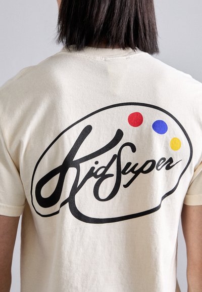 Beige cotton t-shirt featuring a large black "Kid Super" logo on the back, with red, blue, and yellow dots above it. Short sleeves.