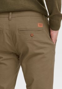 Pantalones caqui de mezcla de algodón, con un bolsillo trasero con botón y una etiqueta de cuero de marca. Textura suave y corte ajustado.