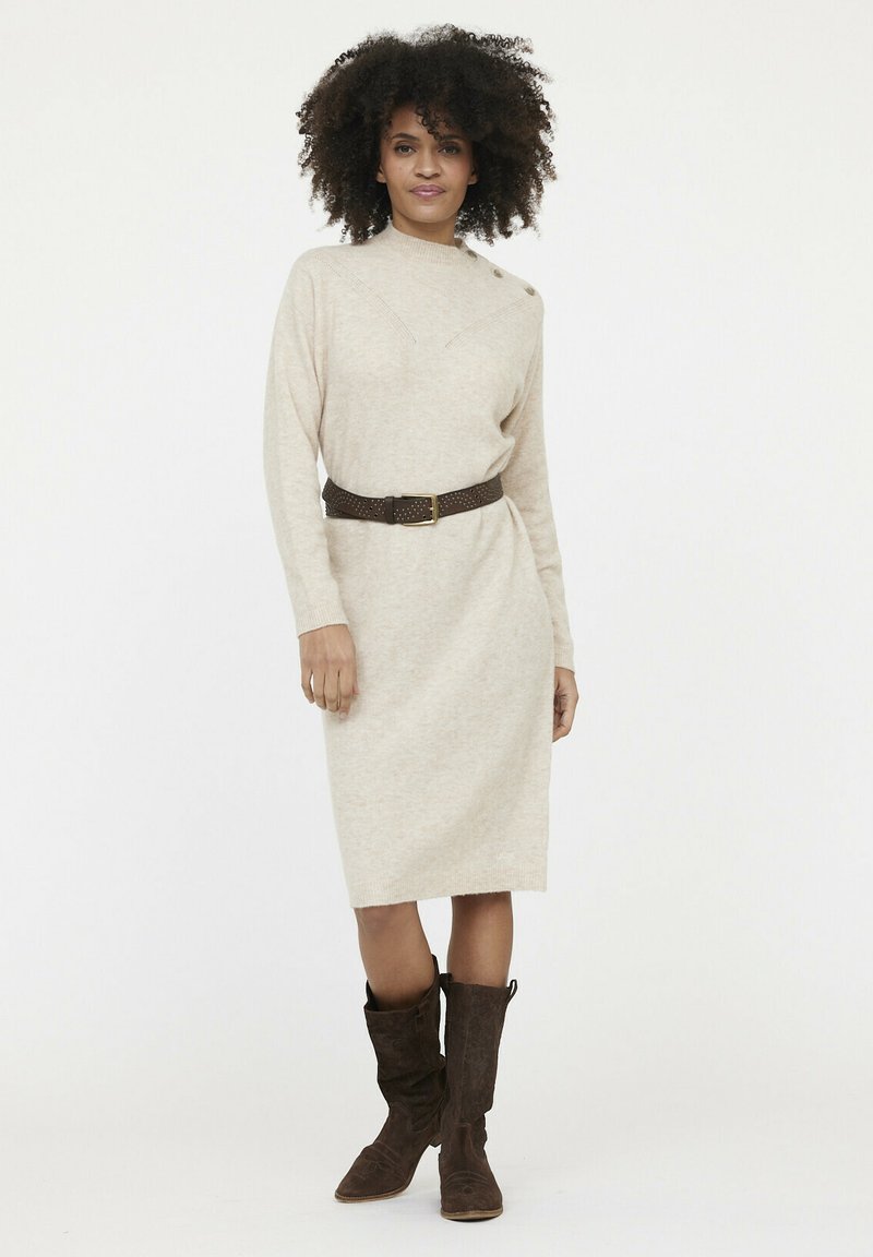 Robe-pull beige à manches longues, col côtelé, détails de boutons décoratifs, cintrée avec une ceinture en cuir texturé marron, portée avec des bottes marron jusqu'aux genoux.