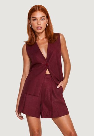 Kvinde med rødt hår iført ærmeløs bordeaux vest og matchende højttaljede shorts, stående med den ene hånd i lommen mod ensfarvet baggrund.