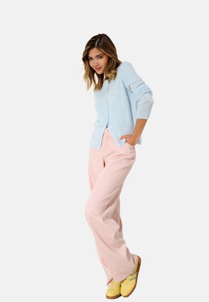 Giovane donna con cardigan azzurro chiaro, pantaloni rosa a gamba larga e sneakers gialle, in piedi con le mani nelle tasche su sfondo bianco.