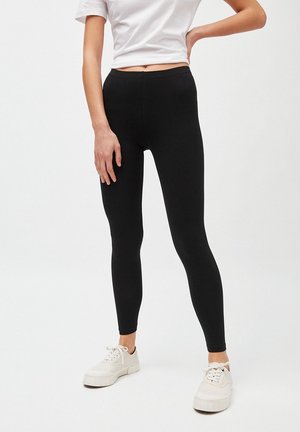 Mujer con leggings negros, camiseta corta blanca y zapatillas blancas, de pie con una mano en la cadera y la otra relajada a un lado.