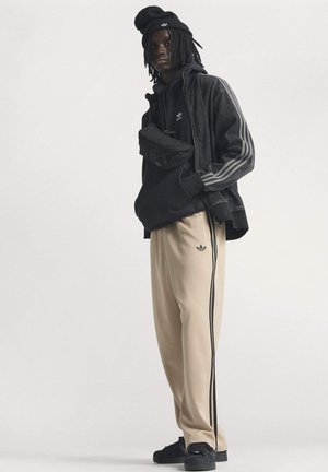 Sort hættetrøje og jakke med grå striber, beige bukser med sorte sidestreger og sorte sneakers, alle med diskrete logoaccenter.