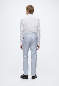 Homme aux cheveux bouclés portant une chemise blanche, un pantalon gris clair et des chaussures marron foncé, debout de dos sur un fond blanc uni.