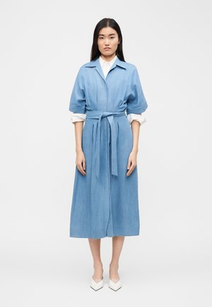 Γυναίκα που στέκεται, φορώντας ένα γαλάζιο μεσαίο φόρεμα με ζώνη και κοντά μανίκια πάνω από μια λευκή μπλούζα μακρυμάνικη και λευκά μυτερά παπούτσια.