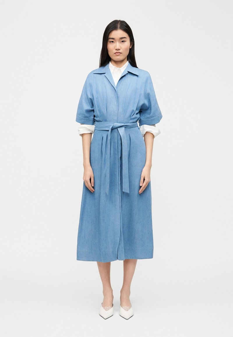 Γυναίκα που στέκεται, φορώντας ένα γαλάζιο μεσαίο φόρεμα με ζώνη και κοντά μανίκια πάνω από μια λευκή μπλούζα μακρυμάνικη και λευκά μυτερά παπούτσια.