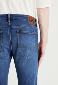 Μπλε τζιν με καφέ δερμάτινη ετικέτα που γράφει «Lee», διαθέτοντας δύο πίσω τσέπες και κίτρινες ραφές. Κλασικό υλικό τζιν.