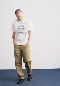 T-shirt blanche à manches courtes avec un logo d'arbre et le texte "Timberland", associé à un pantalon vert olive et des baskets noires, se tenant sur un tapis bleu.