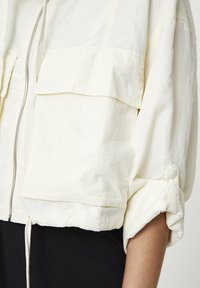 Lekka kremowa kurtka z zamkiem z przodu, dwoma dużymi kieszeniami na piersi oraz podwijanymi rękawami zapinanymi na zatrzaski. Materiał o fakturze, lekko pomarszczony.